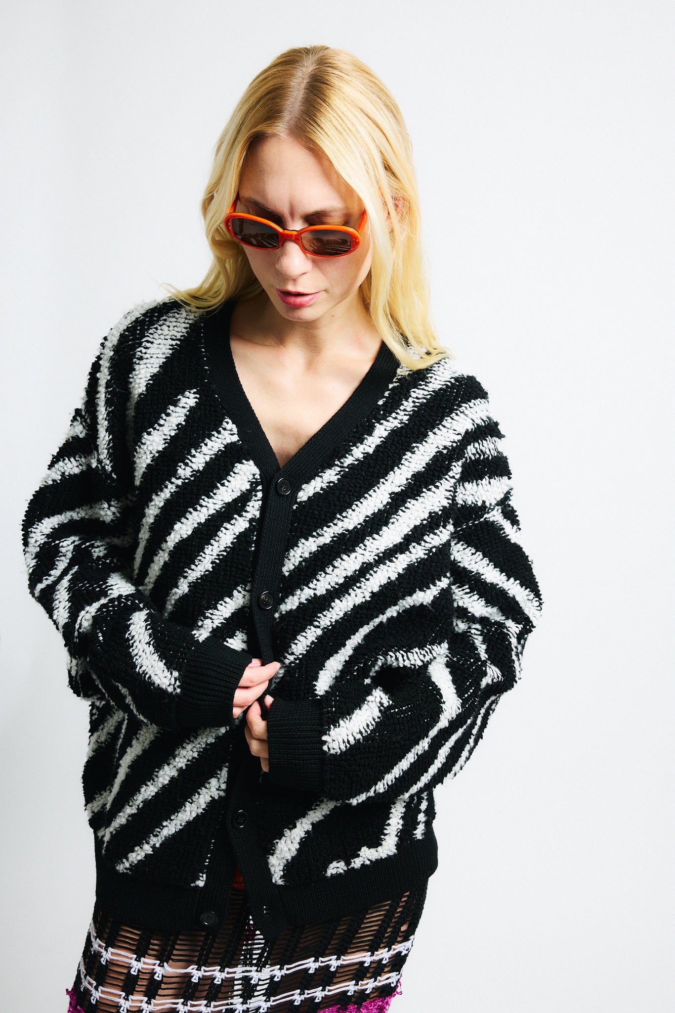 Link to "Christian" cardigan. Image is of a female model wearing "oil spill"/"zebra" print fuzzy v-neck cardigan in black and white stripes with black see-through striped knit dress and orange sunglasses.