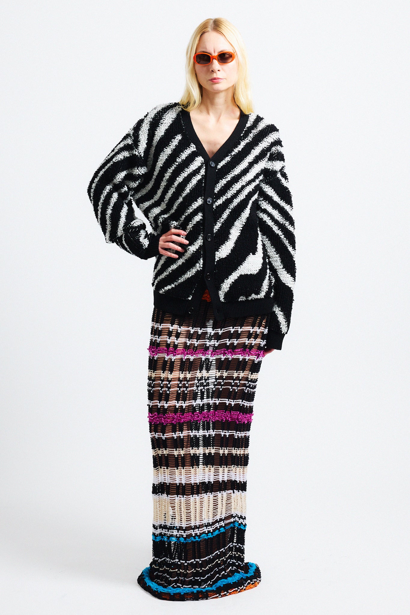 Link to "Christian" cardigan. Image is of a female model wearing "oil spill"/"zebra" print fuzzy v-neck cardigan in black and white stripes with black see-through striped knit dress and orange sunglasses.